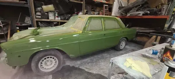 Green vintage car parked in a cluttered garage workshop with shelves and tools nearby