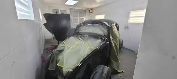 Damaged car in a white garage, wrapped with green protective covering and a smashed rear section.