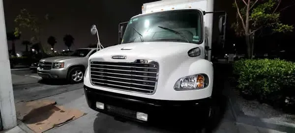 White delivery truck parked at night beside a silver car in a lot with shrubs and buildings