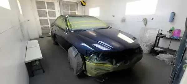 Dark blue car in a small garage with masking tape and covered front bumper, under repair or repainting.