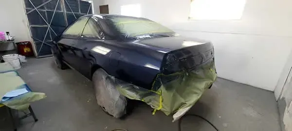 Dark blue sedan masked and taped for paint in a garage, with wheels covered and front bumper removed