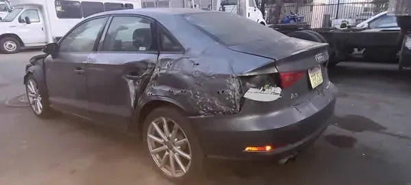 Damaged dark gray sedan with rear-end collision, parked on a wet street
