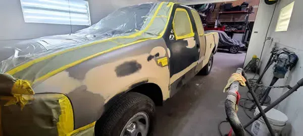 Car in auto body shop with sanded primer patches, yellow masking tape, and repair equipment nearby