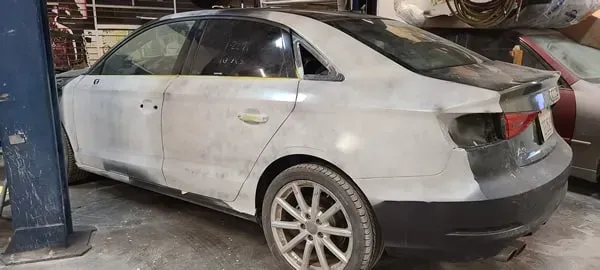 White sedan with dents and scuffs, parked indoors beside a blue car lift in a garage