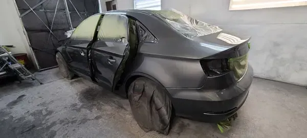 Black sedan with a crushed roof and broken doors, parked inside a garage with debris on the floor