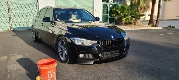 Black sedan parked on a sunny street beside a building, with an orange bucket in front
