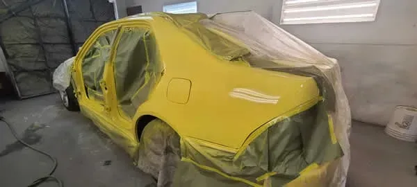 Yellow car covered in masking tape and plastic inside a spray paint booth