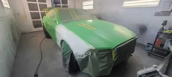 Green car under repair in a paint booth, covered with masking paper and tape.