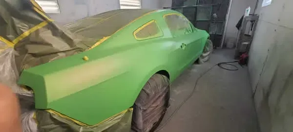 Green car body in a garage, partially masked with tape and paper during painting.