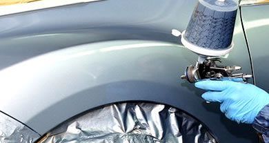 Blue car fender being sprayed with paint by a gloved worker holding a spray gun