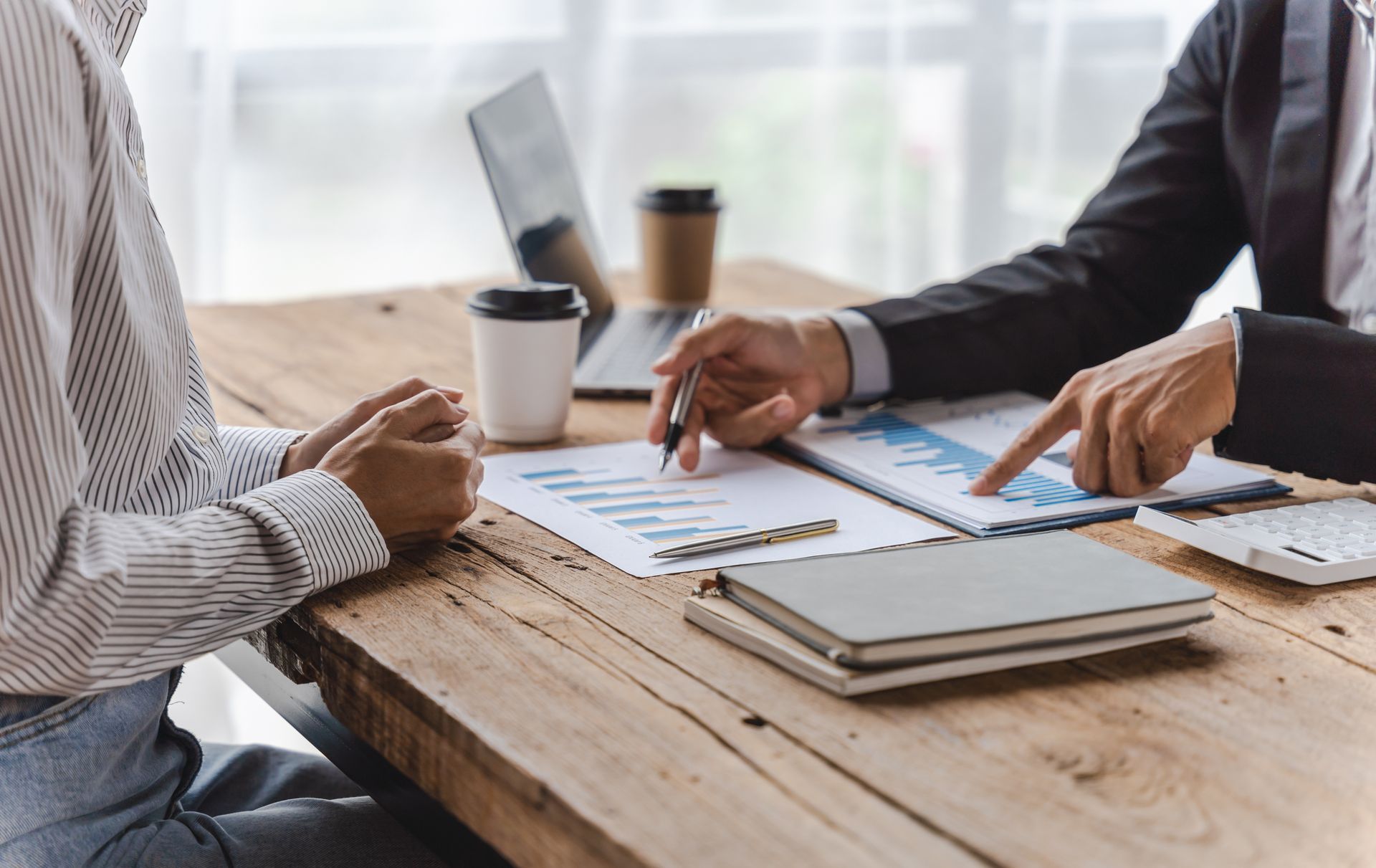 Two people review documents at a wooden table with coffee cups and a laptop in a bright office.