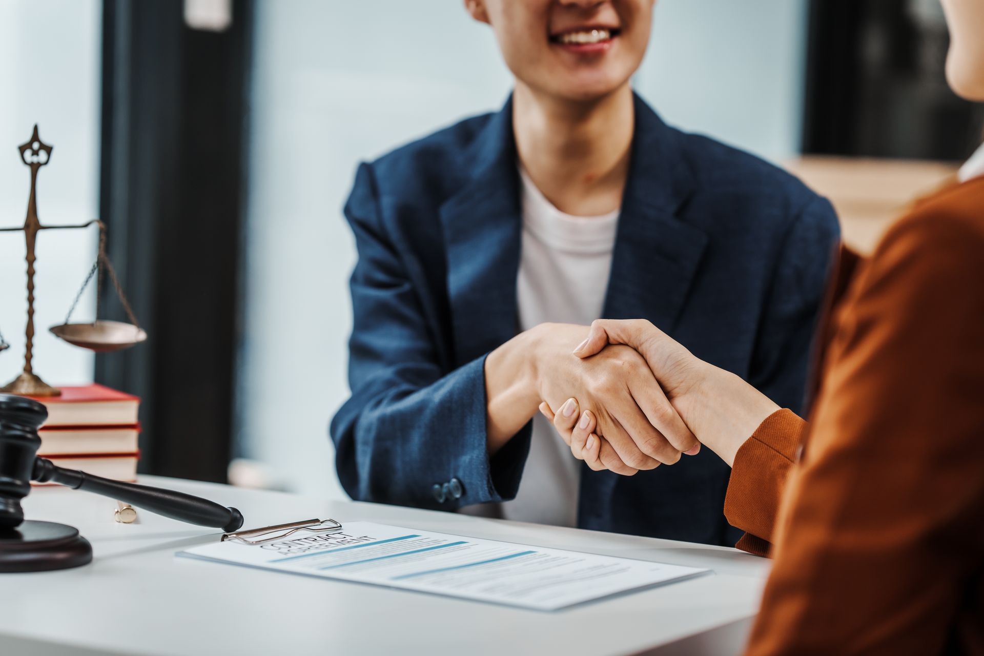Two people shaking hands across a desk with documents and a scale in a professional office setting