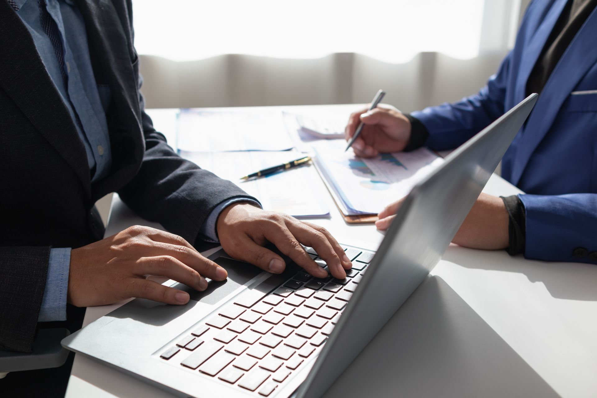 Two people in business attire working at a laptop during a meeting, with notes on a desk