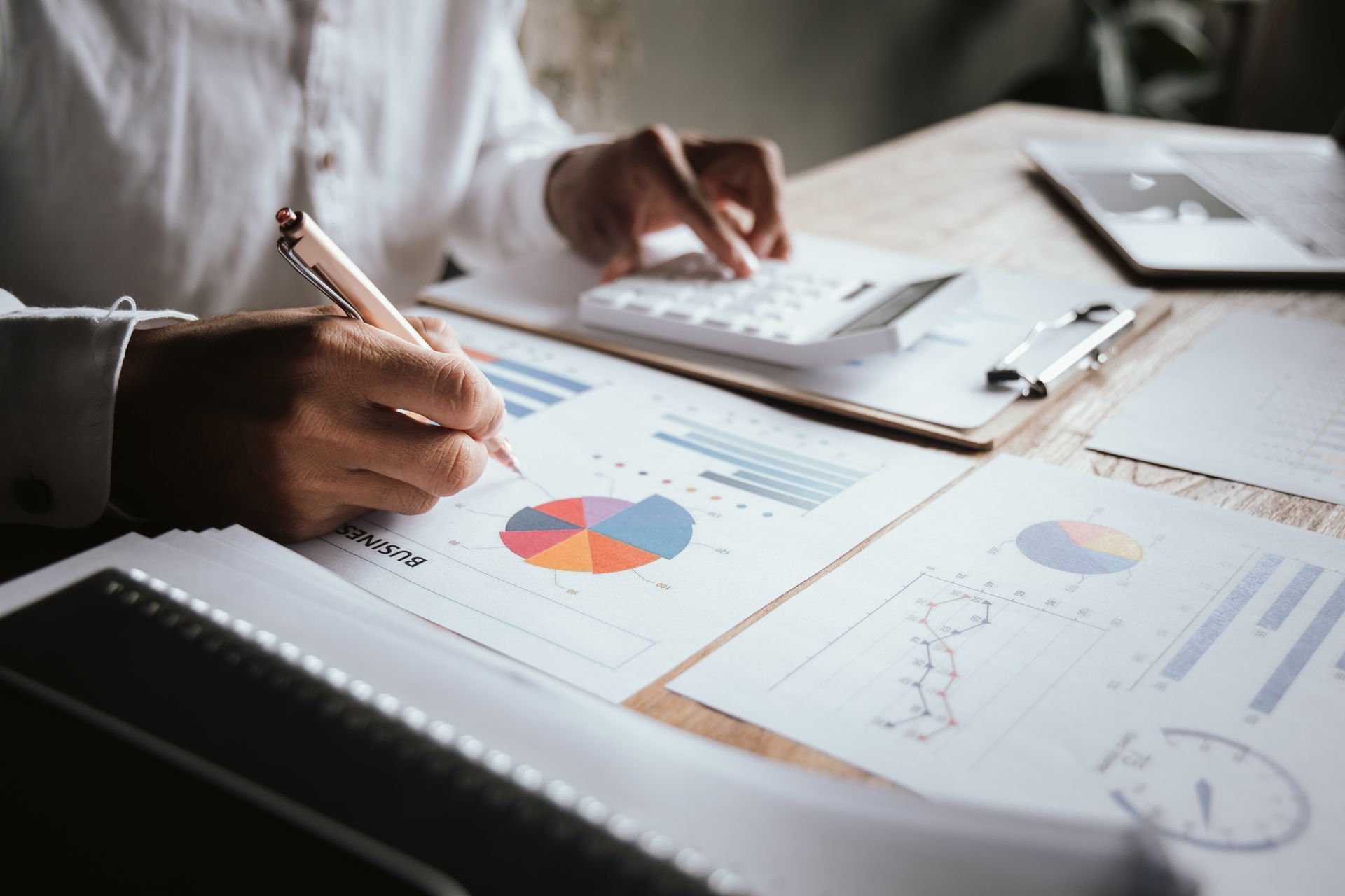 Hands writing notes beside printed charts and a laptop on a desk