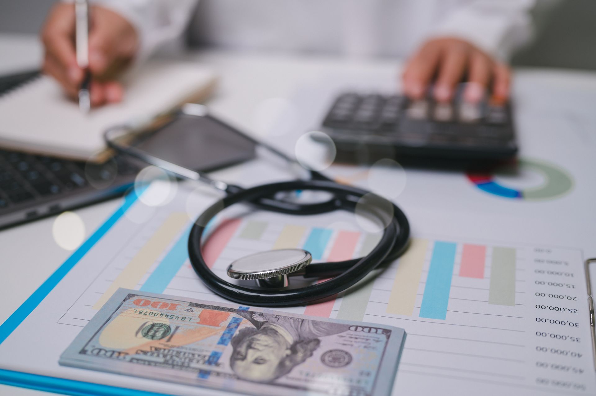 Hands using calculator over financial charts with a stethoscope and banknote on desk