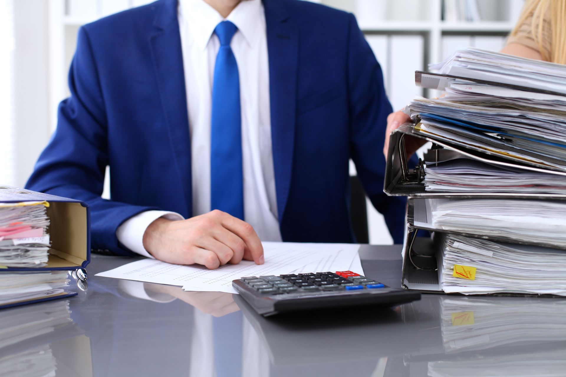 Businessperson in blue suit with calculator and stacked paperwork at a desk