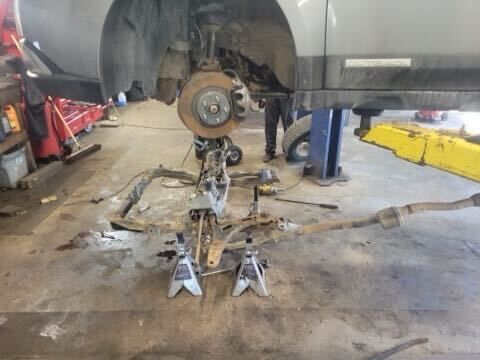 A car is elevated on a lift in an auto shop with its subframe and suspension components removed and supported by jack stands.