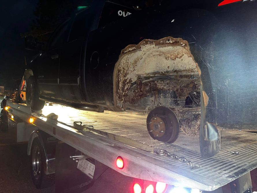 A black pickup truck with severe rust in the rear wheel well, secured on the bed of a tow truck at night.