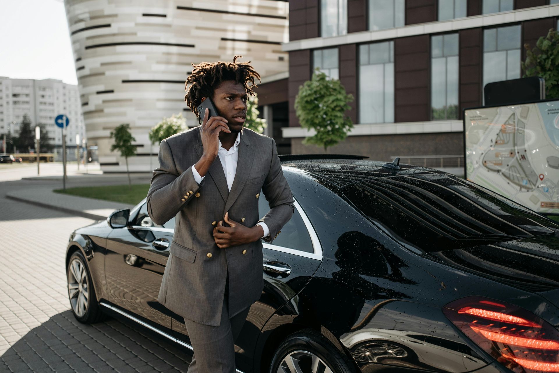 Man in gray suit talking on phone beside a black sports car in a modern urban setting