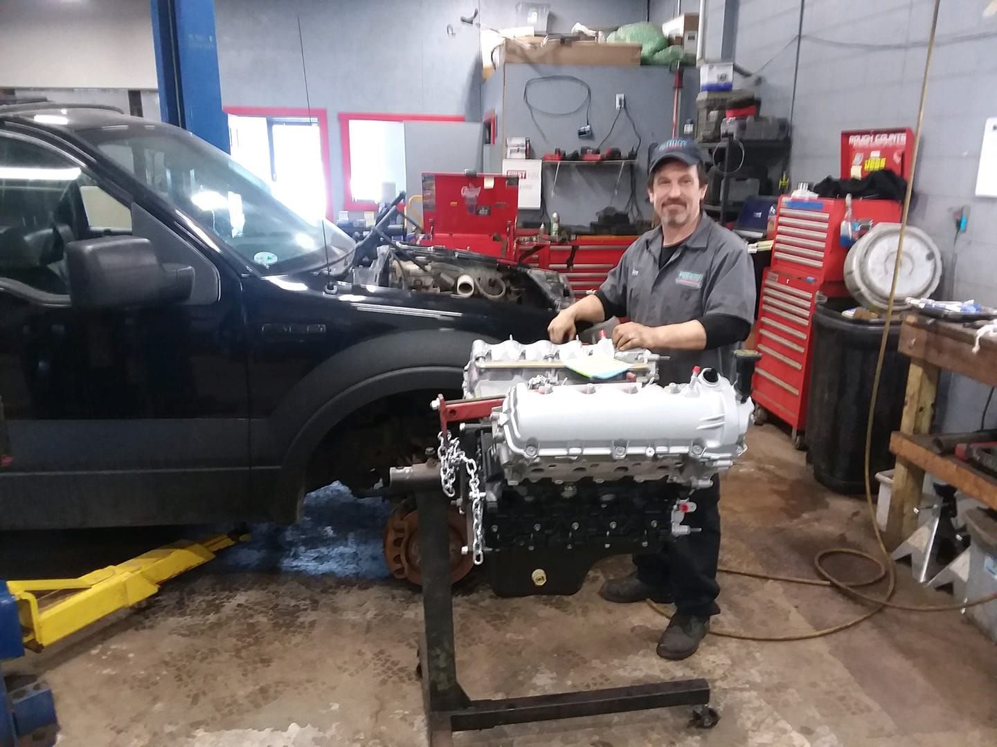 A mechanic smiles while standing next to a newly rebuilt engine on a stand in a busy automotive repair shop.