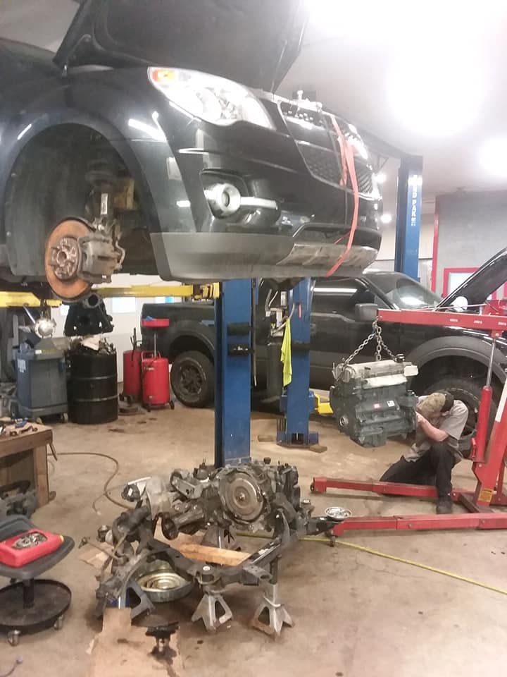 A mechanic works on a car engine in a repair shop with a vehicle lifted on a hydraulic hoist and an engine crane nearby.