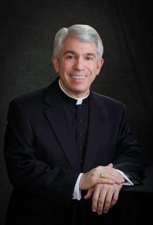 Portrait of a smiling Catholic priest in a black clerical suit against a dark background.
