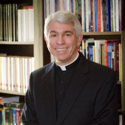 A smiling priest in a black clerical shirt stands in front of bookshelves.