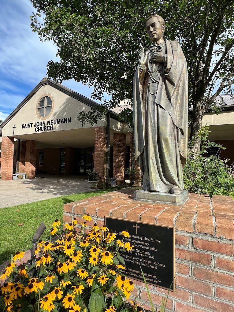 Statue of a robed figure by a library entrance, with yellow flowers and a brick sign under a tree.