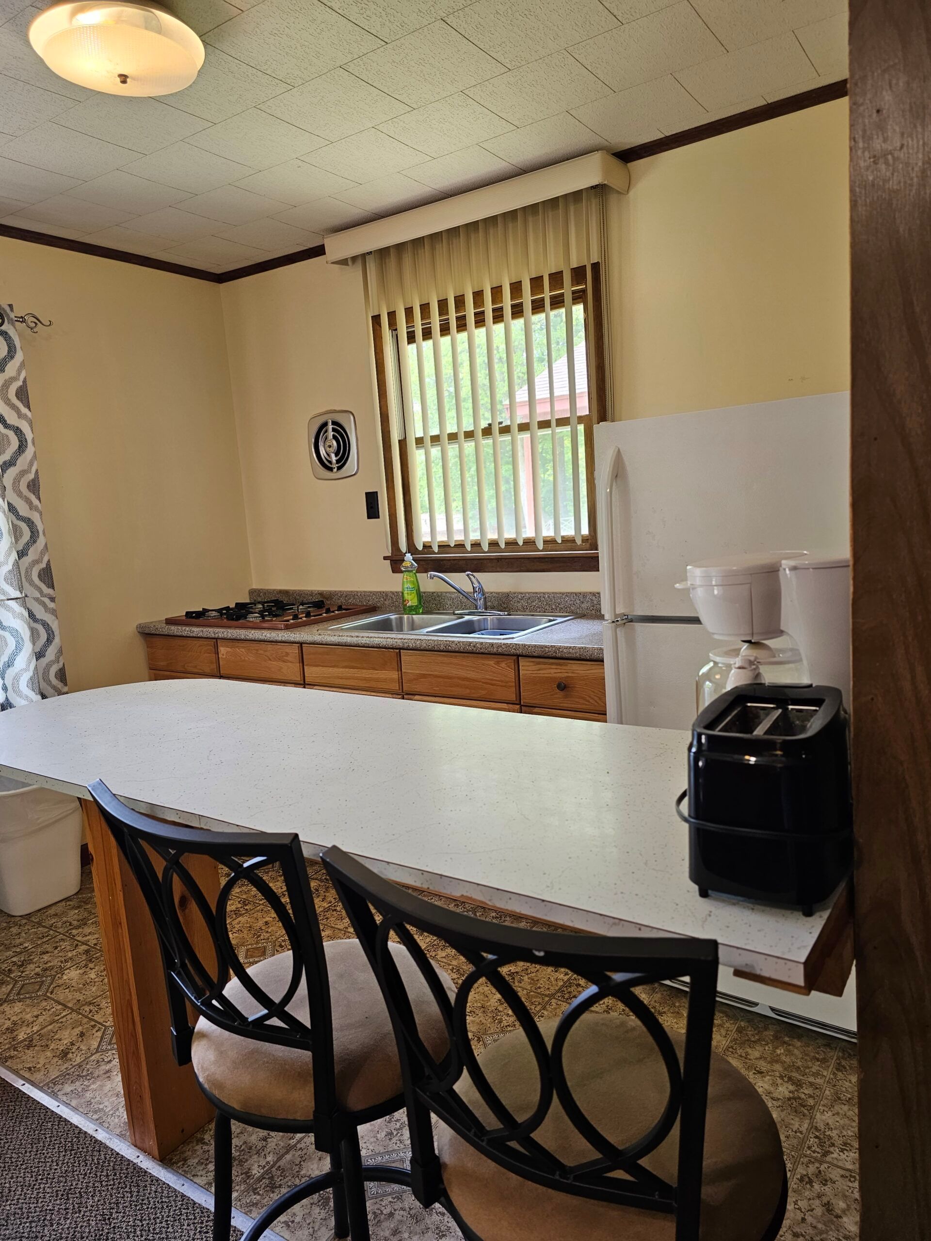 Small kitchen with beige walls, white counter, sink under a window, and two black bar stools in the foreground