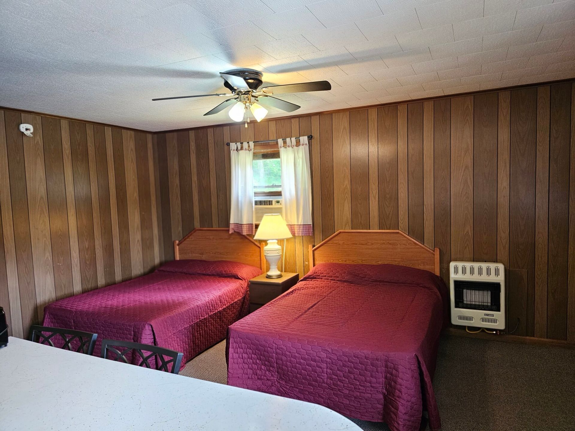 Motel room with two beds, wood-paneled walls, a ceiling fan, and a lamp-lit window.