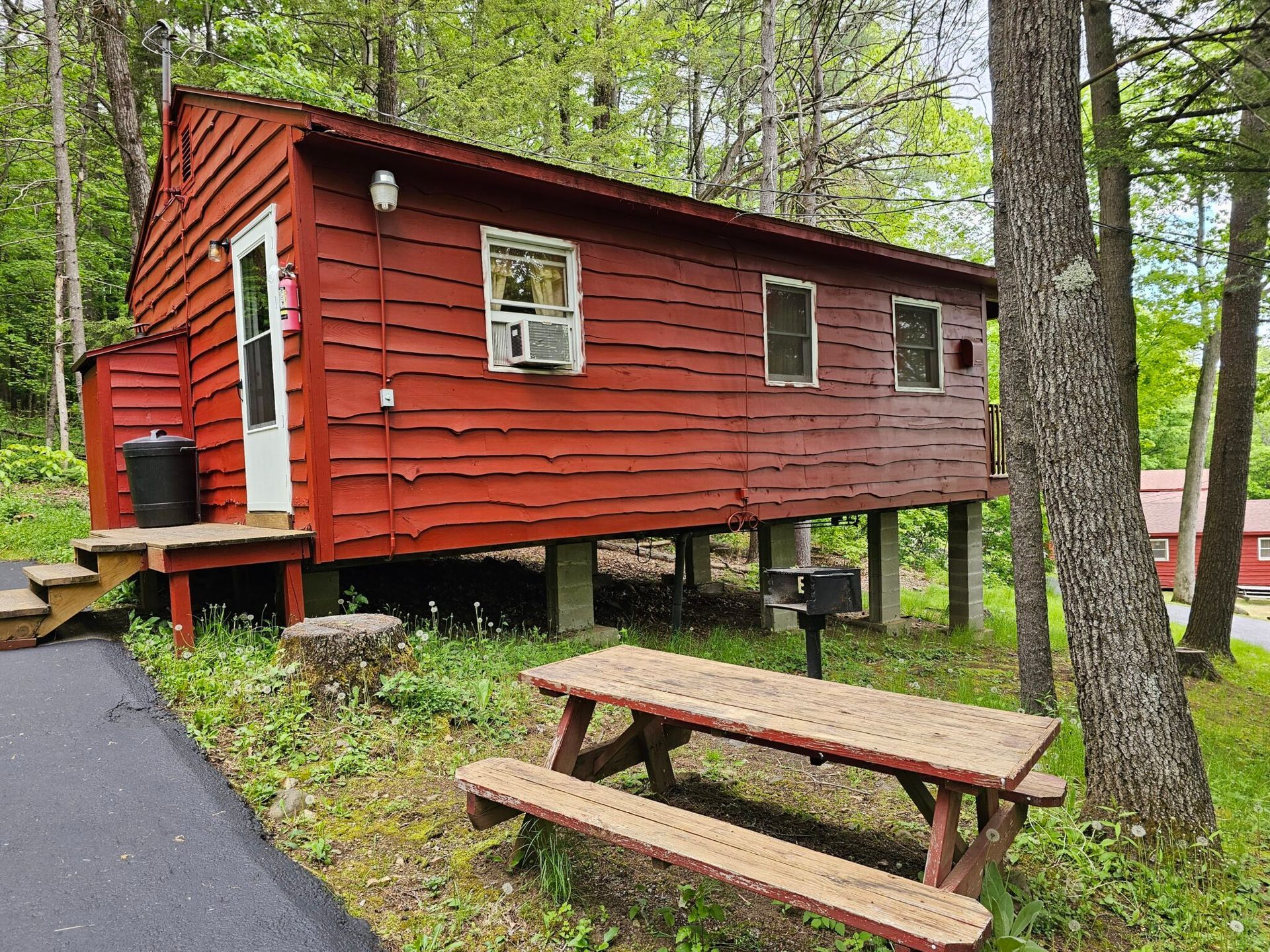 Red cabin in a wooded campsite with a picnic table and paved path nearby