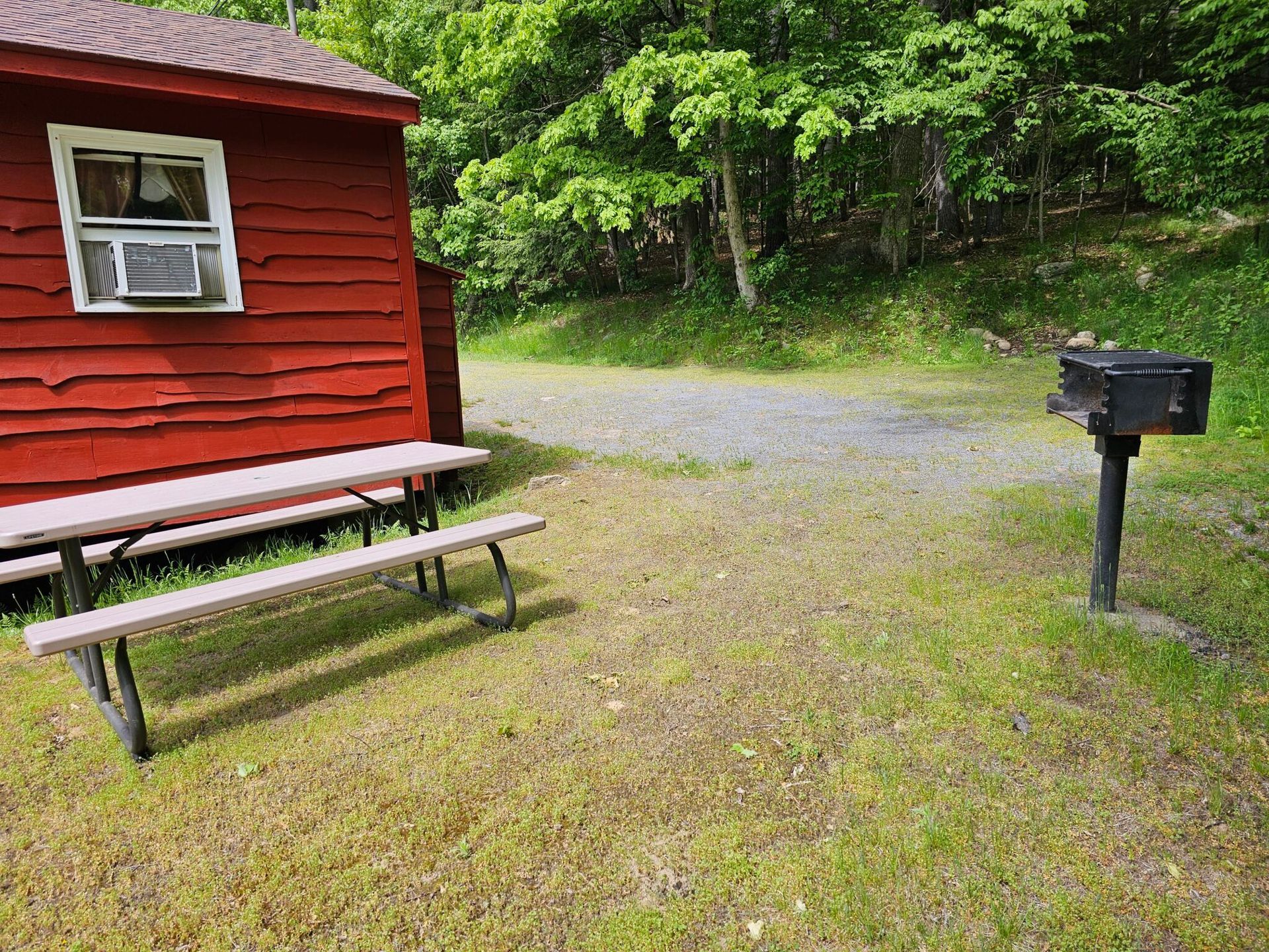 Red cabin with picnic table and black mailbox beside a gravel driveway in a wooded area