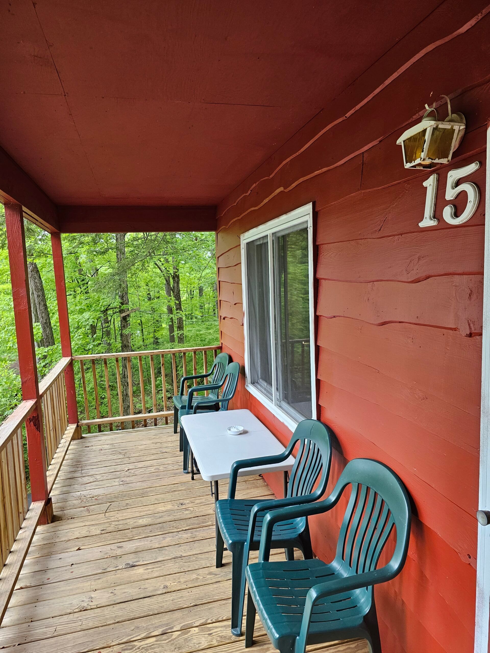 Covered red cabin porch with two blue chairs, a white table, and a wooded view through the railing.