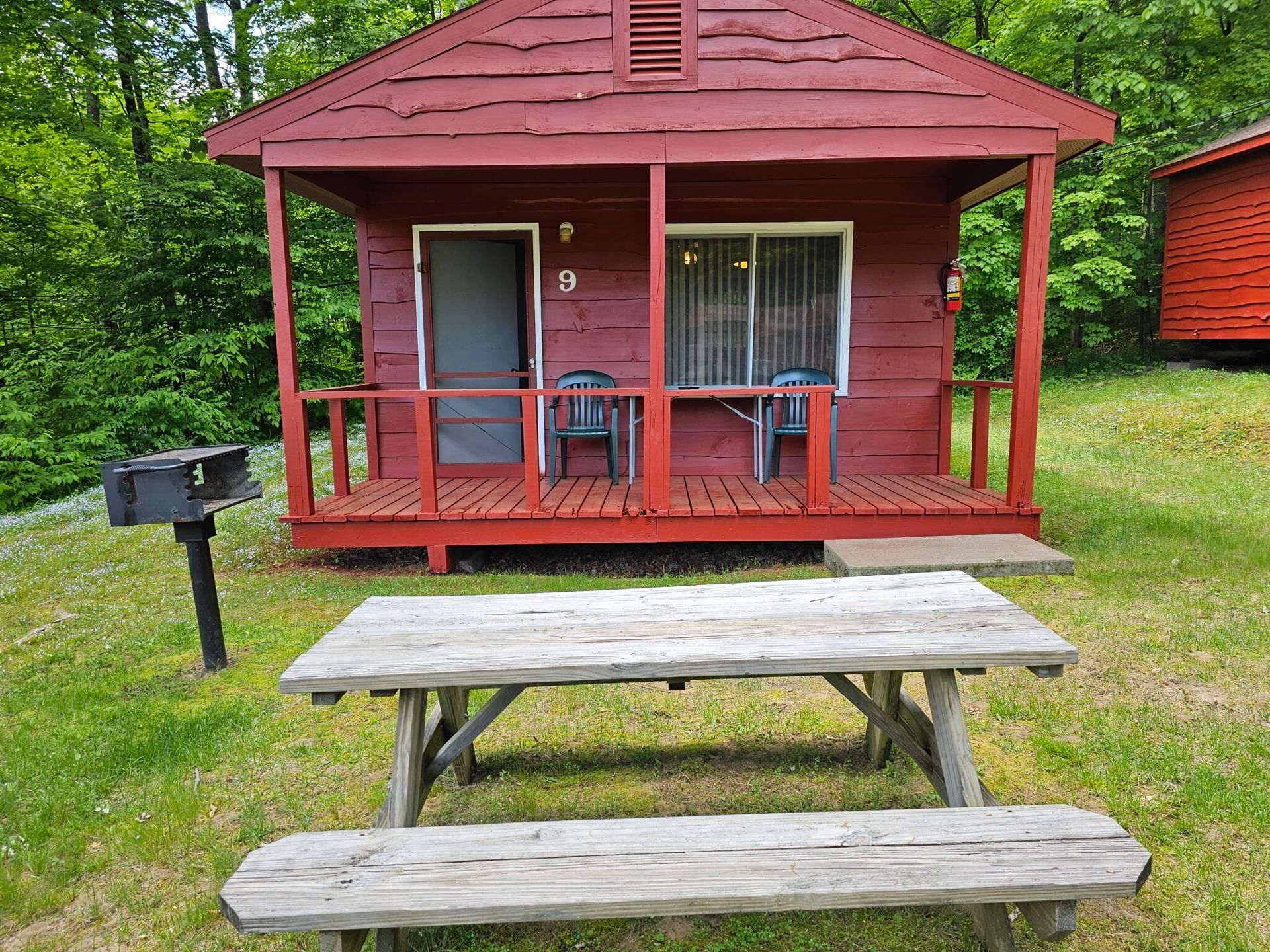 Red cabin porch with chairs, a picnic table in front, and a black grill on the left.