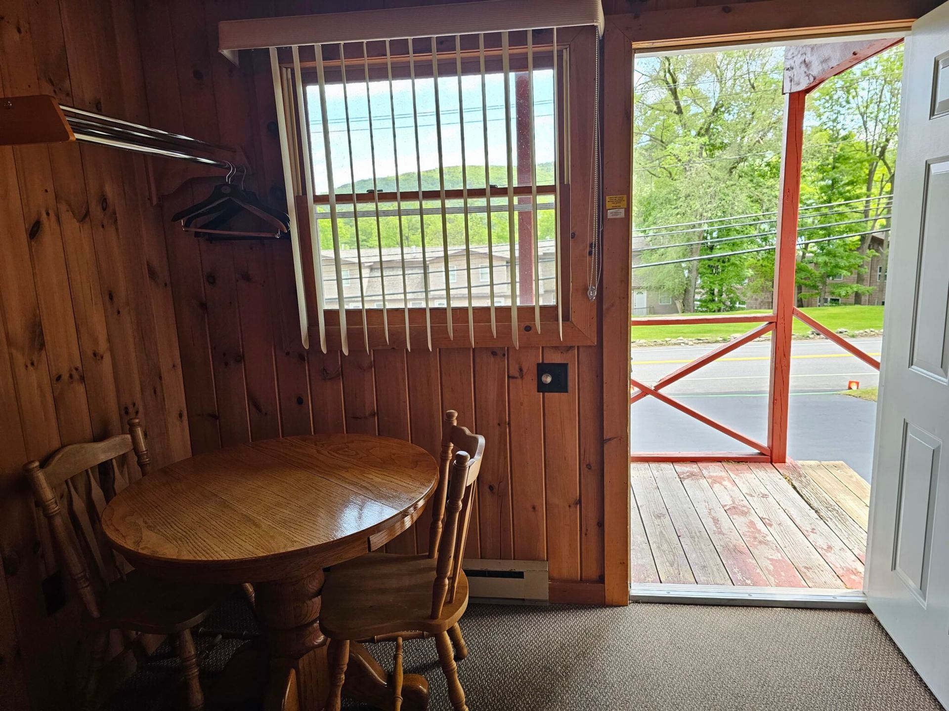 Wood-paneled room with round table and chair, looking out through an open doorway to a porch and road outside.