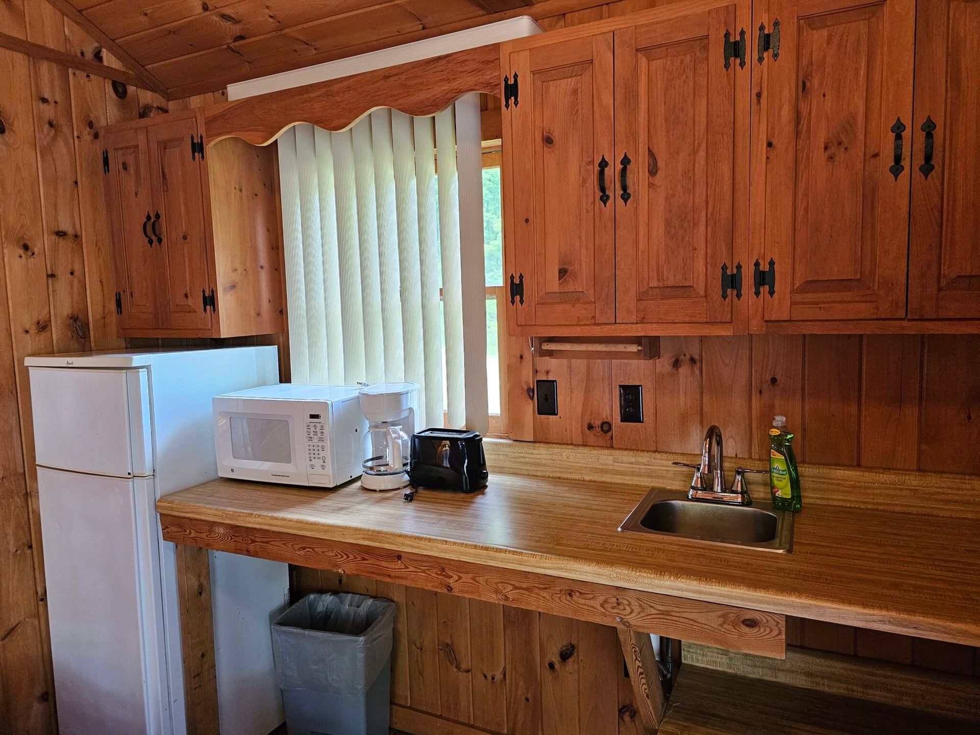 Wood-paneled kitchen with a microwave, coffee maker, sink, and white refrigerator under a curtained window