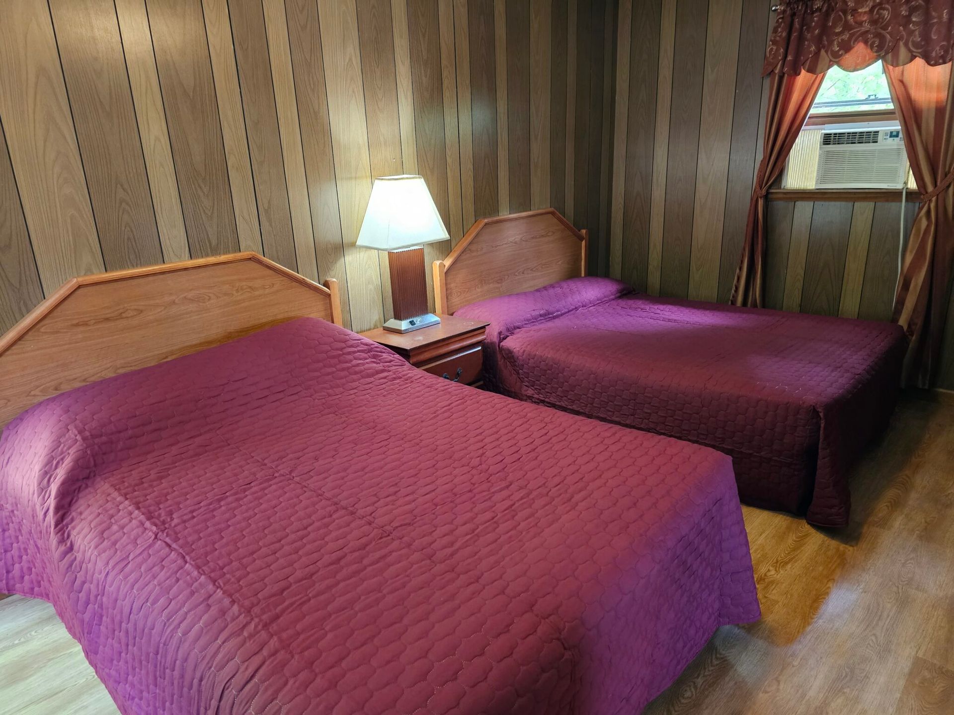 Two twin beds with purple quilts in a wood-paneled motel room, lit by a bedside lamp and window.