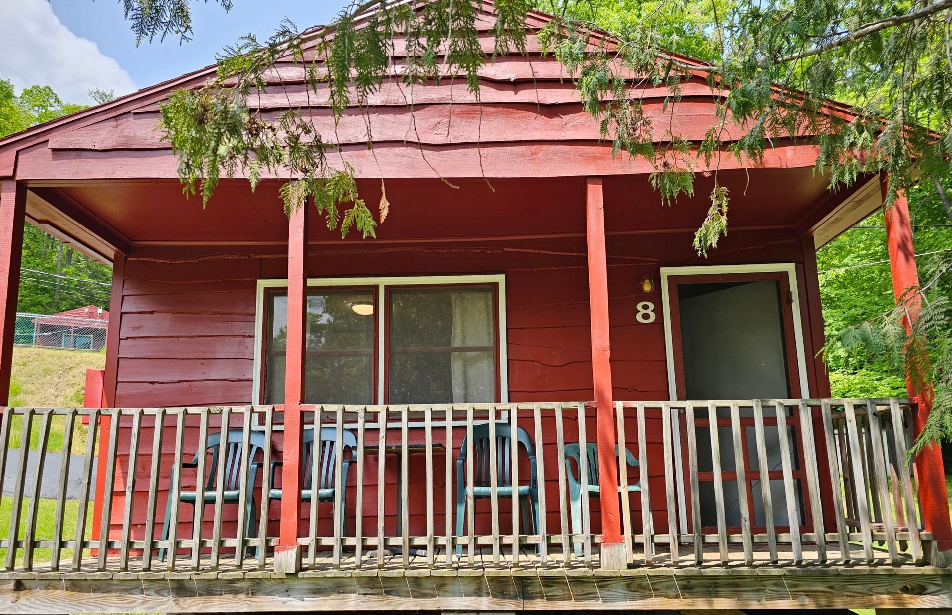 Red wooden house with a porch, white railing, and a sliding glass door under leafy tree branches