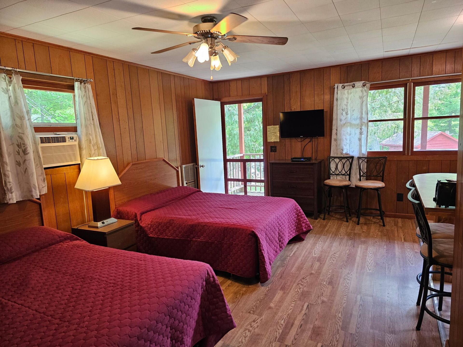 Cozy wood-paneled bedroom with two red beds, ceiling fan, TV, and windows looking outside