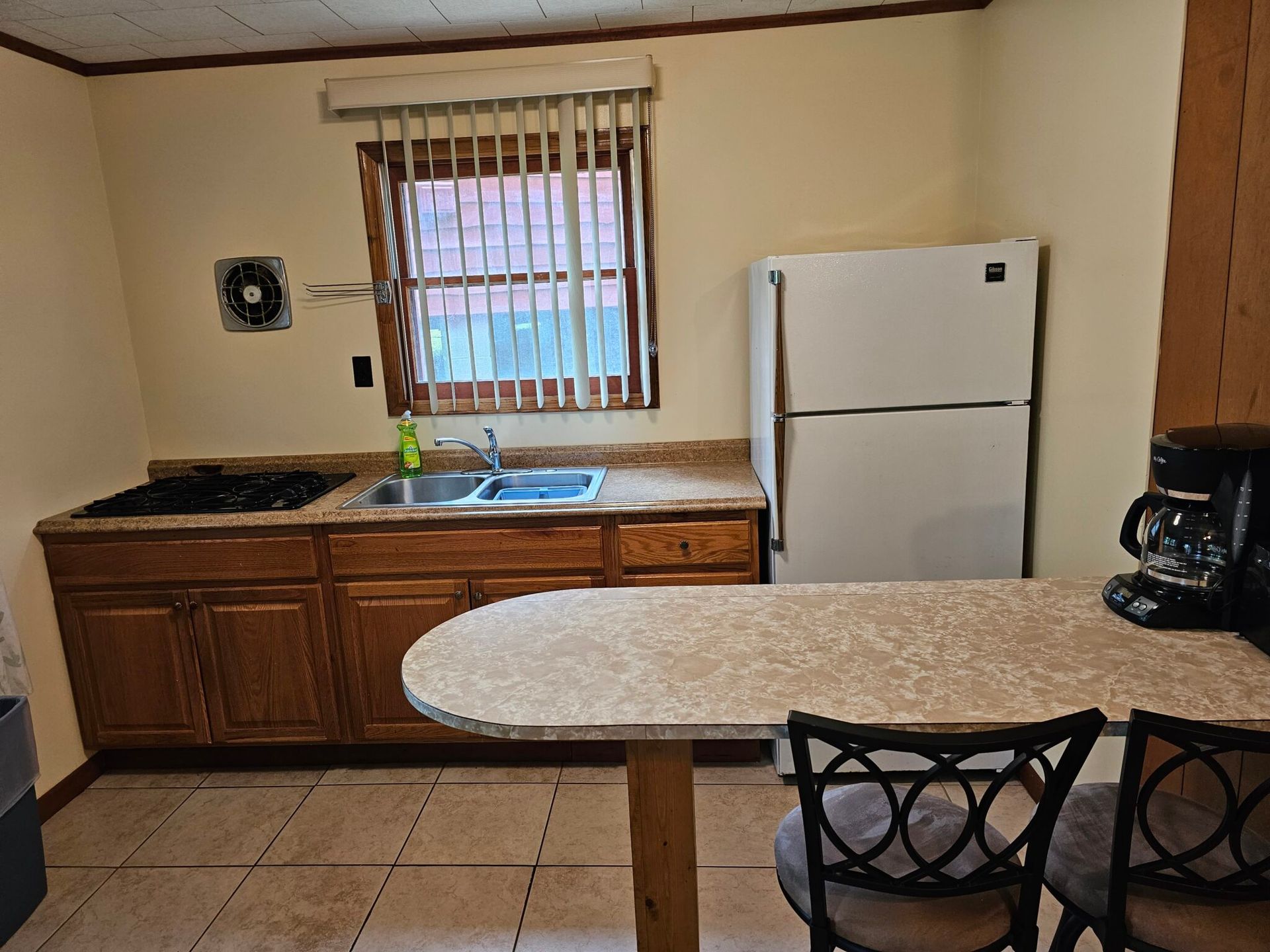 Small kitchen with wooden cabinets, granite island, sink under window, white fridge, and coffee maker.