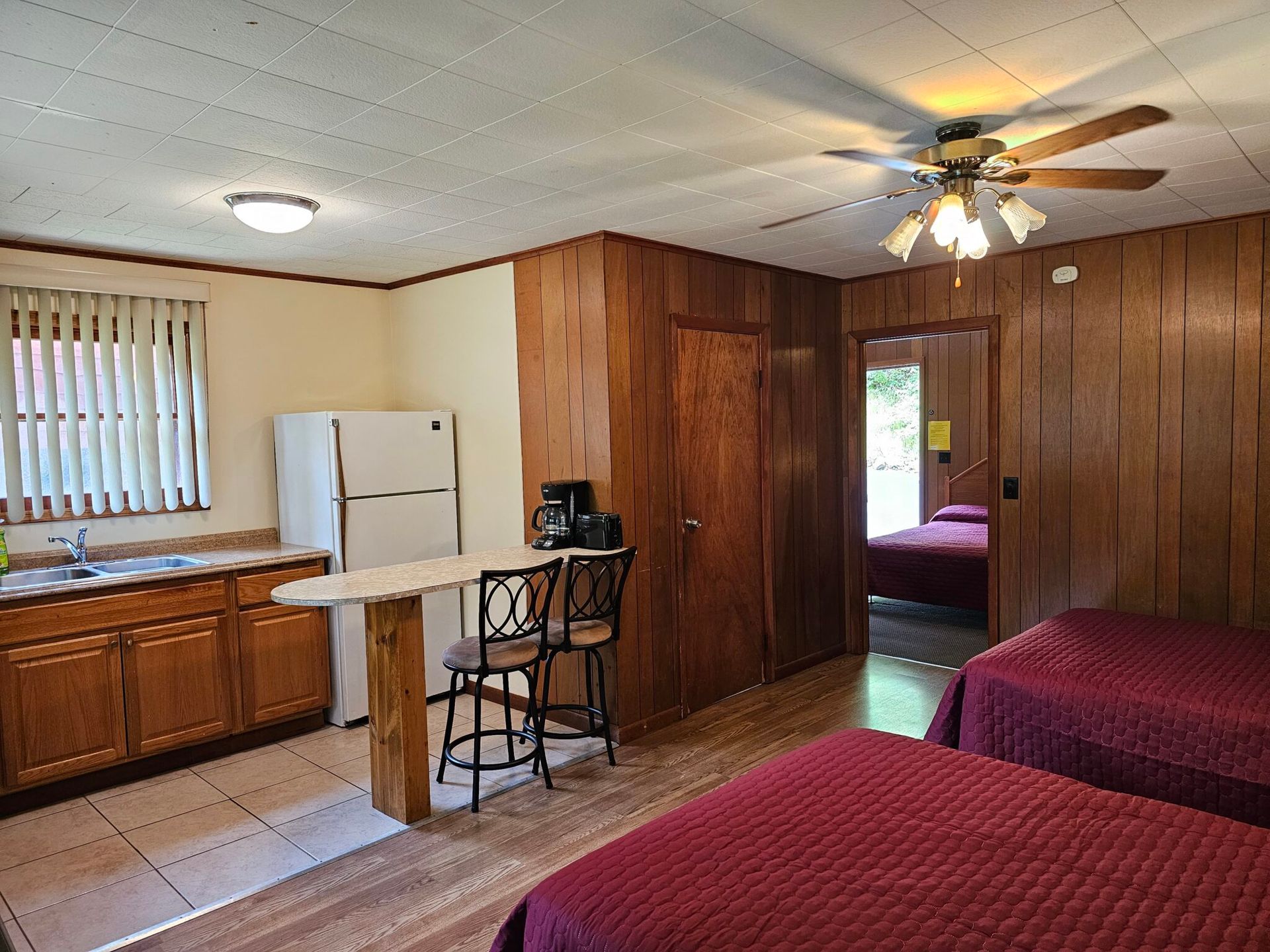 Cozy wood-paneled studio with kitchenette, two beds, ceiling fan, and doorway to bedroom