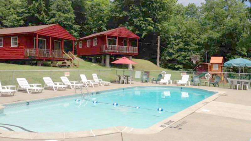 Outdoor swimming pool with lounge chairs and red cabins in a wooded resort setting