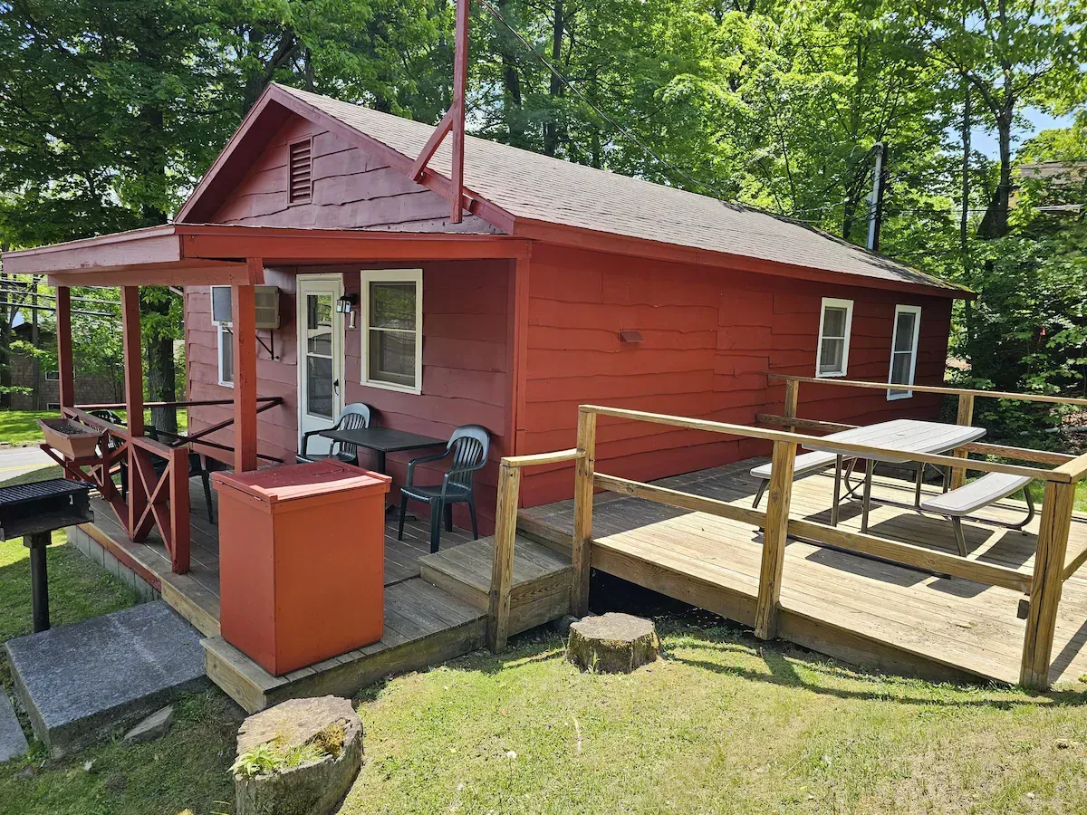 Red cabin with porch and ramp deck in a wooded yard, with outdoor chairs and a red grill.
