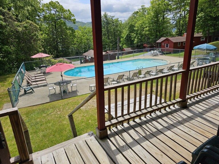 Covered deck overlooking outdoor pool with umbrellas, lounge chairs, and trees on a sunny day