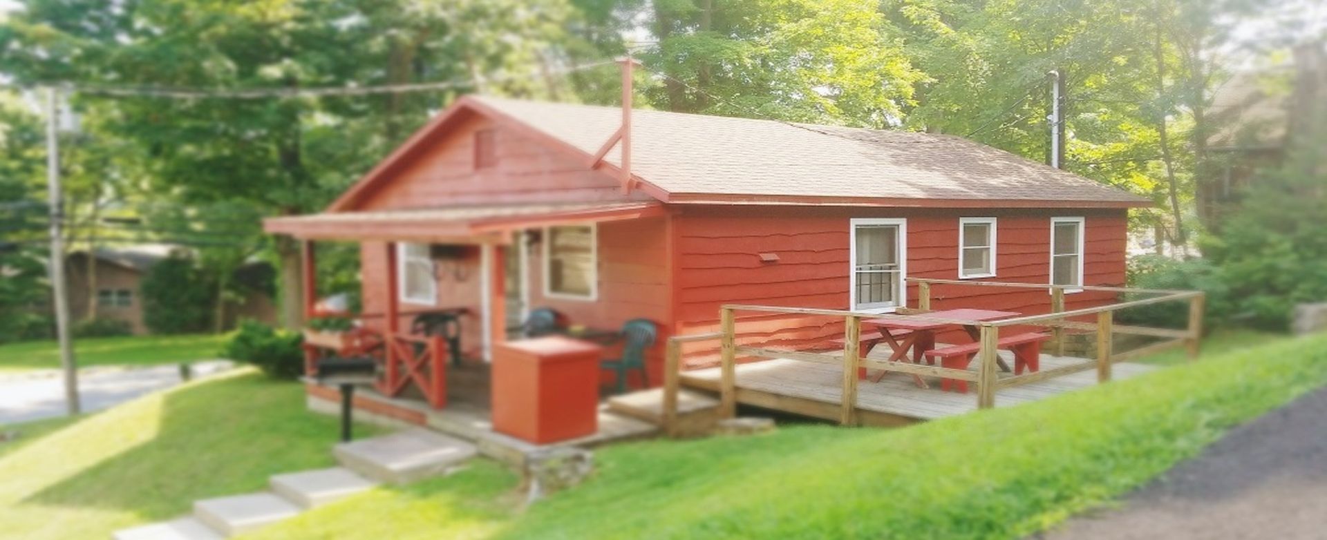Red cabin-style house with porch and red roof on a grassy lot, surrounded by trees and a paved road