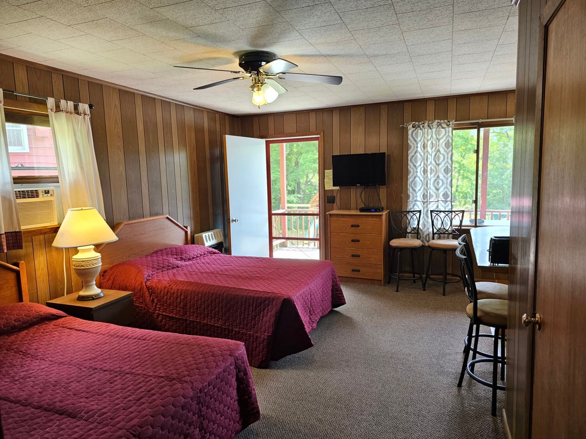 Bedroom with two beds, wood-paneled walls, TV, desk, and a sliding glass door to a balcony.