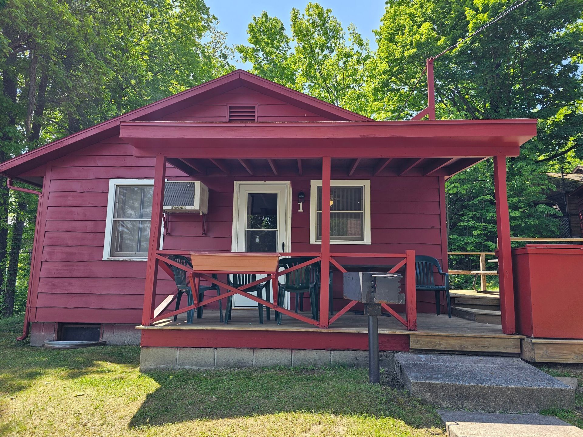 Small red cabin with a front porch, white-trimmed windows, and a wooded background