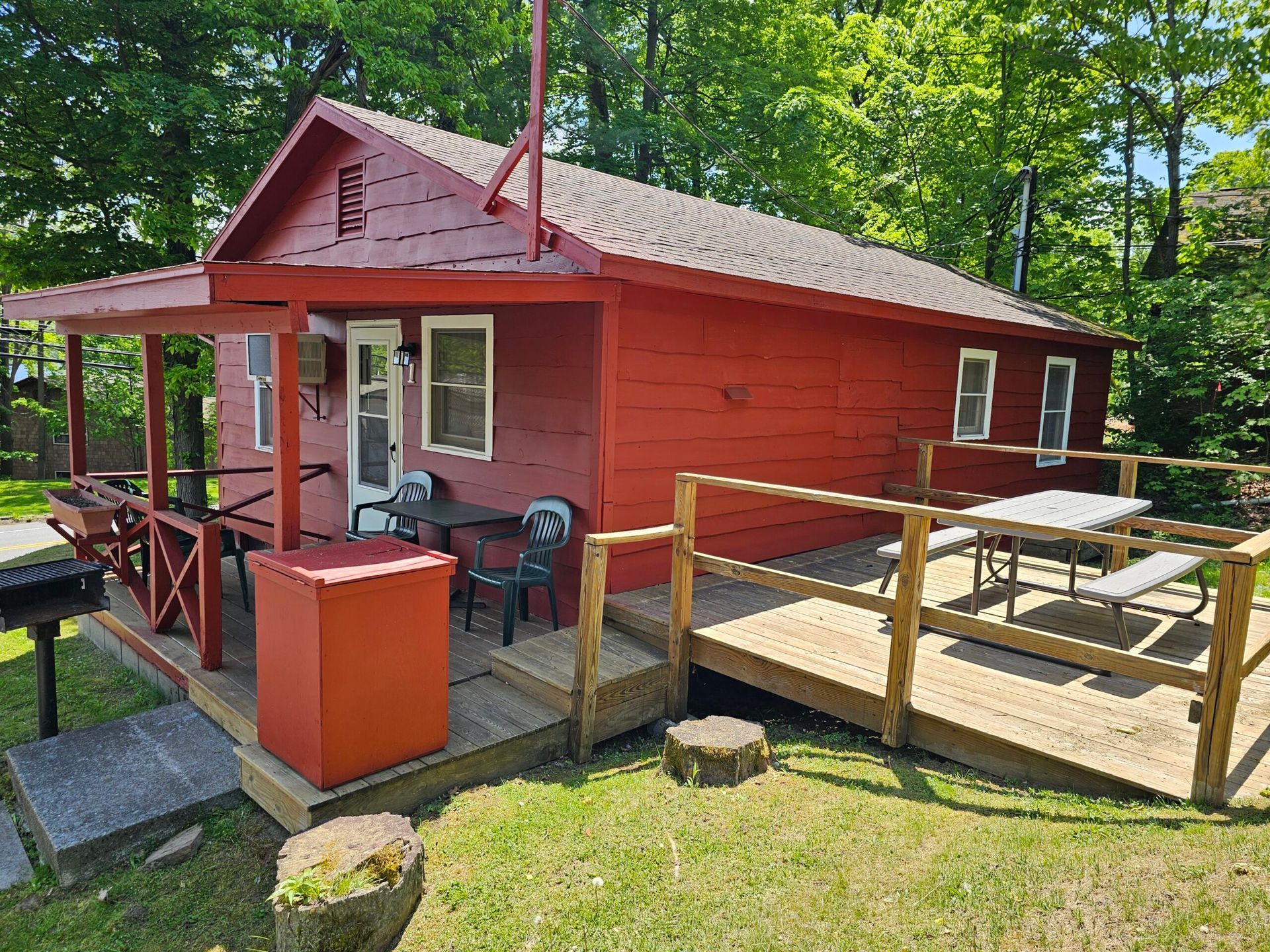 Red cabin with porch, ramp, and wooden deck surrounded by trees and grass