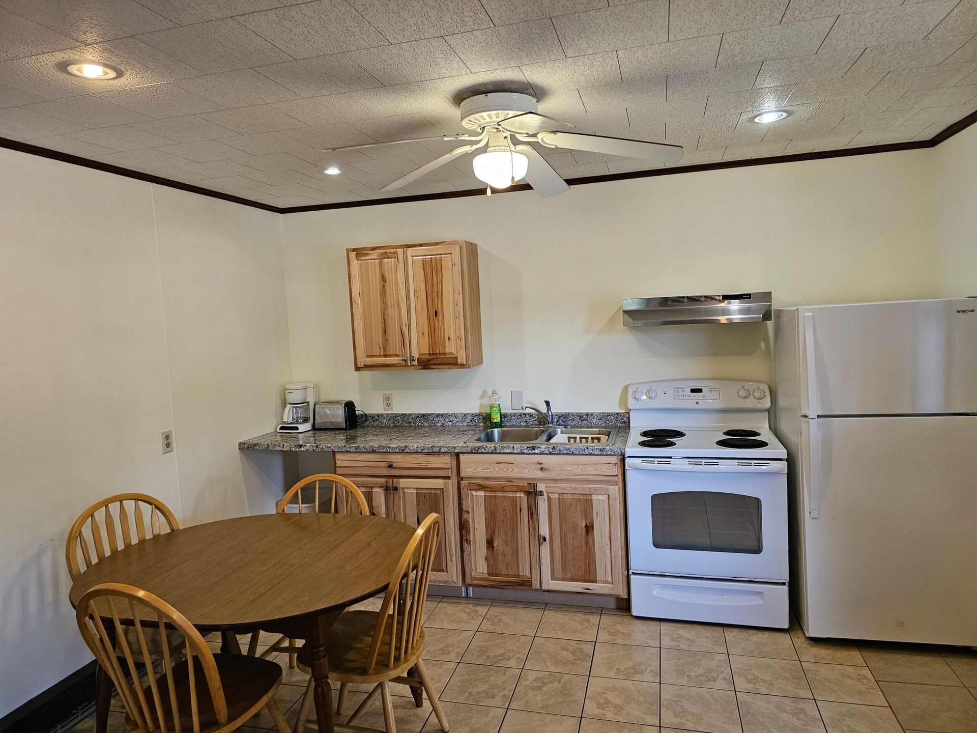 Small kitchen with wooden table and chairs, white stove and fridge, and ceiling fan.