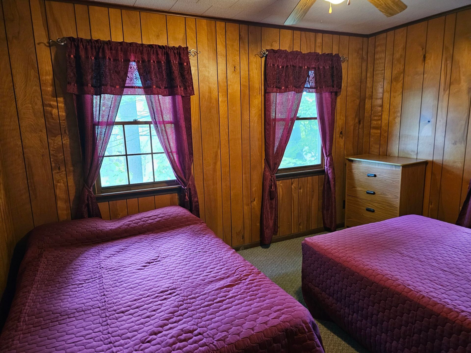 Wood-paneled bedroom with two beds, red bedspreads, burgundy curtains, and a dresser by the window